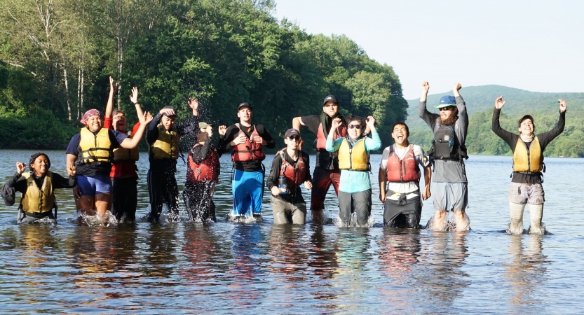 A group of people wearing lifejackets stand in knee-deep water and lift their hands in celebration. 